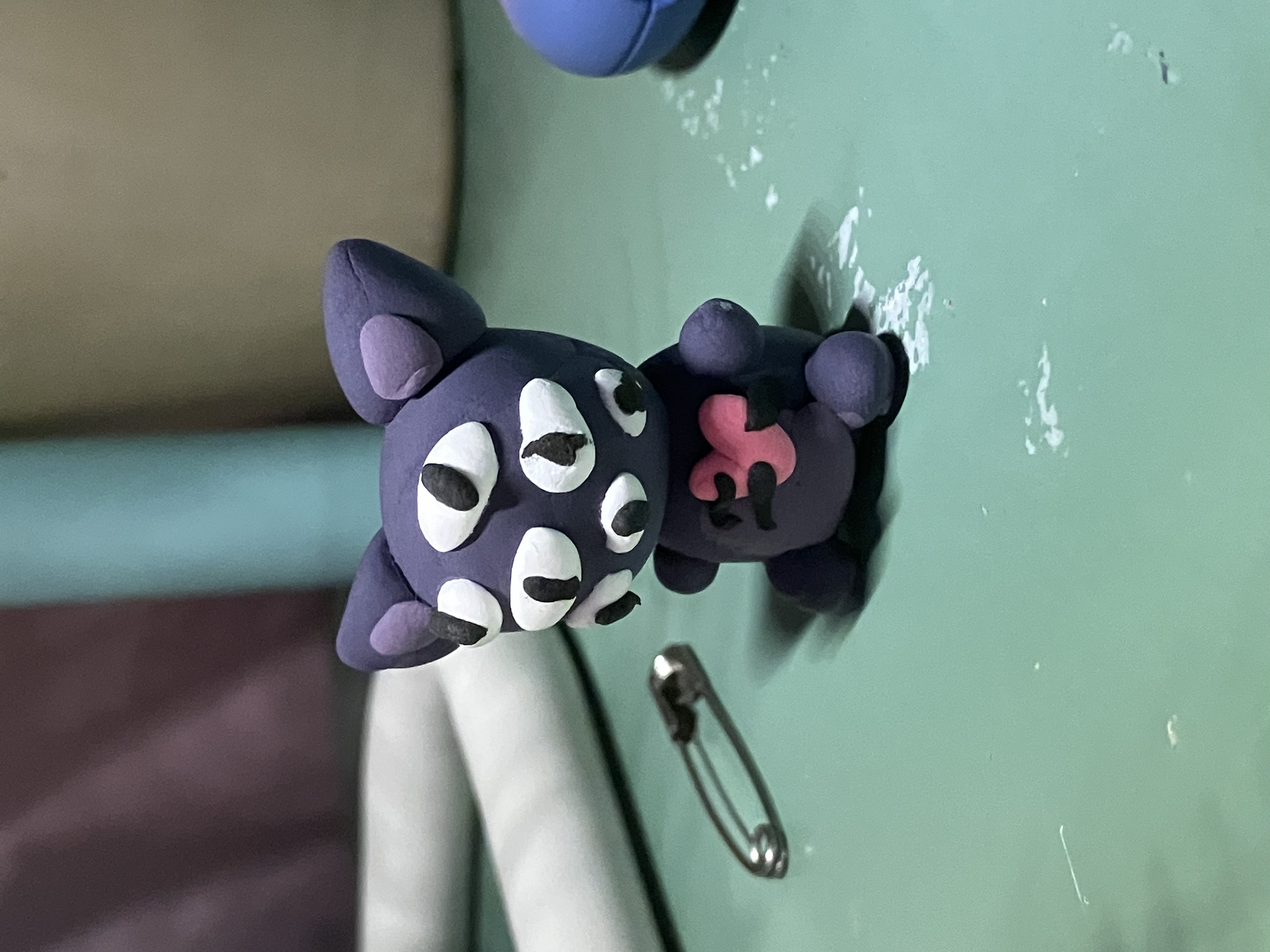 A clay many-eyed purple cat with a pink heart sewn to its chest.